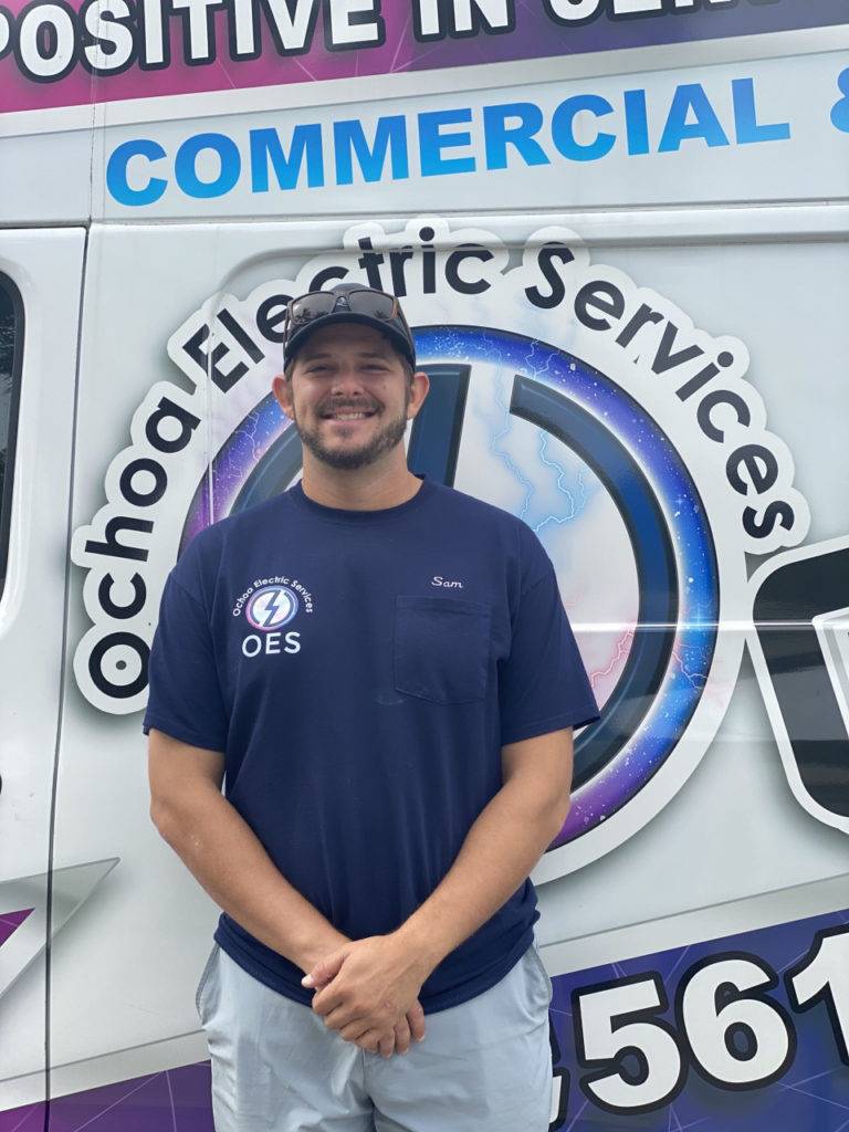 Ochoa Electric Services official van with contact details and "45-hour service" wording on it, with man in cap and dark blue tshirt and shorts standing beside the van